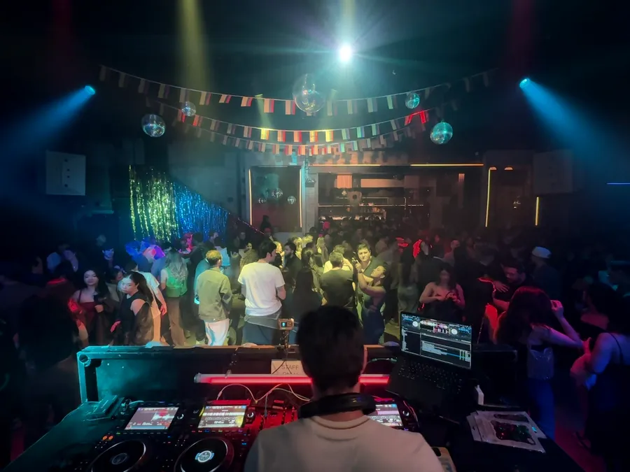 View from the DJ booth over a packed dance floor with disco balls and string lights overhead